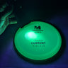 A Meridian Discs Current disc golf disc glowing bright green under UV light, with a hand holding a black UV lamp on the left.